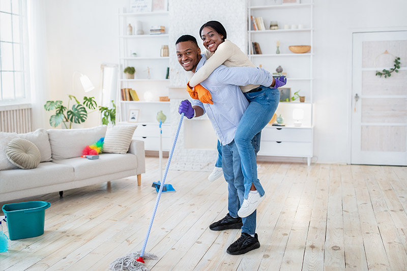 Couple cleaning the floors of their home with mop while woman is on man's back