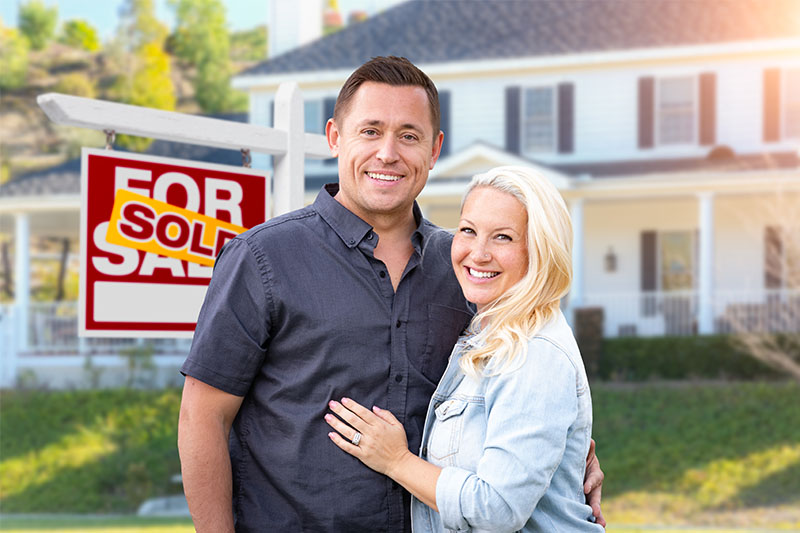 Happy Couple Standing In Front Of House With For Sale Sign